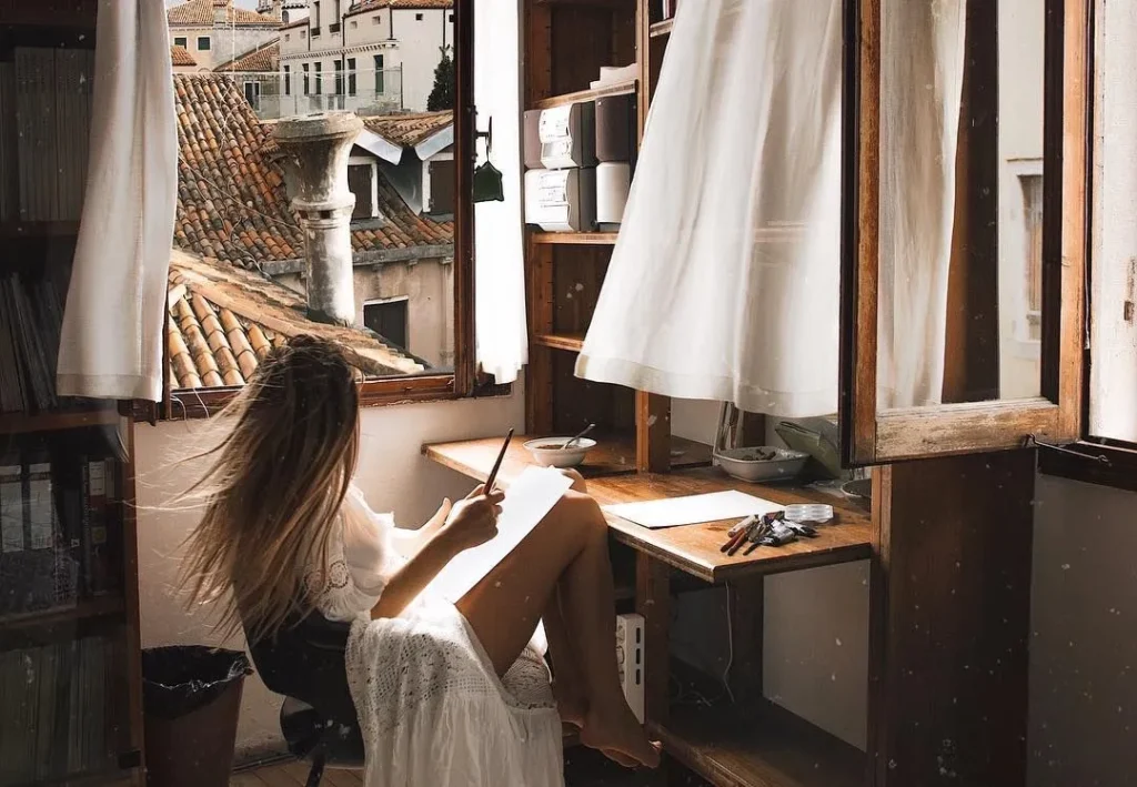 A thoughtful woman gazing out of an open window, holding a pen and paper, seeking inspiration or contemplating her thoughts for writing.